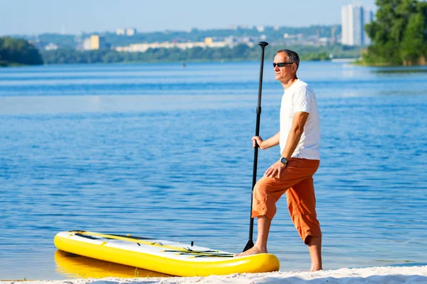 Yetişkin erkek güneş gözlüğü Sup kurulu cityscape arka plan üzerinde sahilde yanında duruyor. Yukarı raket yatılı - stand tatil sırasında harika Aktif istirahat.