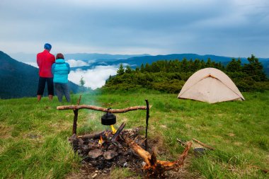 Maceracılar kampı yeşil çayır ateşin ortasına yanında rahatlayın ve dağ sıralarının siste fırtınadan sonra hayranım. Epik seyahat dağlarda. Arkadan görünüm.