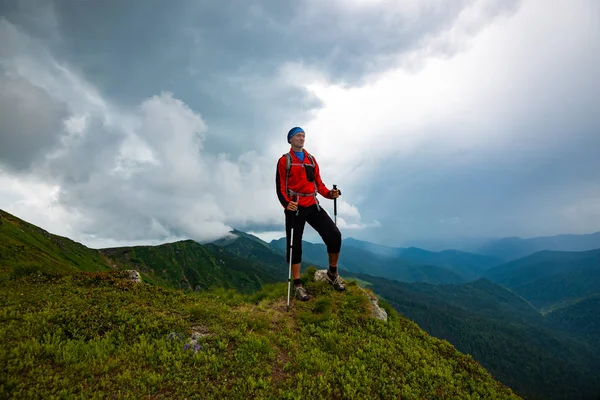 Adventurer is standing on the cliff on the background of low heavy clouds floating above green ridges stretching to the horizon - in mountains before storm. Wide angle.