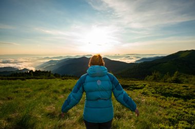 Gezgin, Erkek yeşil dağ yamacında duruyor ve günbatımı sırasında ufuk bulutlu gökyüzü altında uzanan ormanlık tepeler hayran. Arkadan görünüm, arka ışık, geniş açı.   