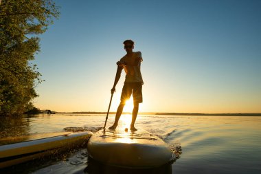 Adam bir Sup kurulu yükselen güneş ışınlarının içinde yola çıkacak. Yukarı raket yatılı - stand doğada awesome Aktif istirahat. Arka ışık.