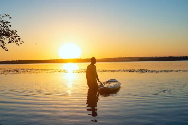 Man relaxes next to a SUP board in a large river and admires sunrise. Stand up paddle boarding - awesome active recreation in nature.