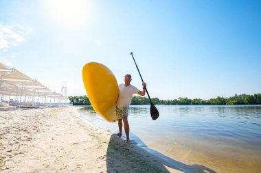 Neşeli adam sahilde elinde Sup kurulu ile ayakta ve kameraya bakıyor. Yukarı raket yatılı - stand harika Aktif istirahat. Geniş açı, arkadan.