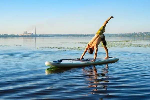 Man practicing yoga on a SUP board during sunrise on a large river. Stand up paddle boarding - awesome active recreation in nature.