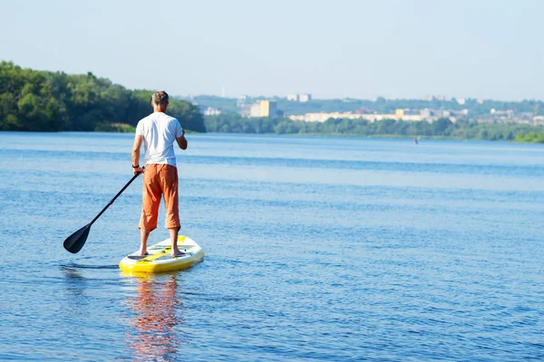 Man sails on a SUP board in large river on the cityscape background. Stand up paddle boarding - awesome active recreation. Back view.