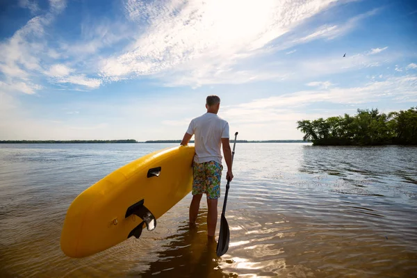 Man is standing with a SUP board and a paddle in his hands on the beach against blue sky background. Stand up paddle boarding - awesome active recreation. Back view, wide angle.