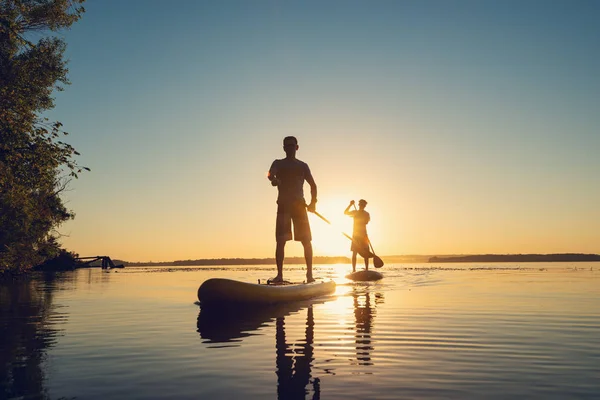 Men, friends sail on a SUP boards in a rays of rising sun. Stand up paddle boarding - awesome active recreation in nature. Backlight.