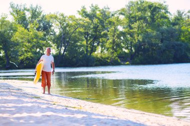Adam bir Sup kurulu ile plaj boyunca elinde yeşil ağaçlar arka plan üzerinde bir güneşli sabah yürüyor. Yukarı raket yatılı - stand harika etkin açık rekreasyon. 