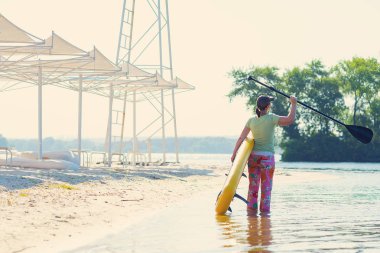 Mutlu kadın sahilde elinde Sup kurulu ile ayakta ve harika sabah hayran. Yukarı raket yatılı - stand harika etkin açık rekreasyon. Arkadan görünüm.