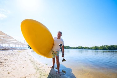 Neşeli adam sahilde elinde Sup kurulu ile ayakta ve kameraya bakıyor. Yukarı raket yatılı - stand harika etkin açık rekreasyon. Geniş açı, arkadan.