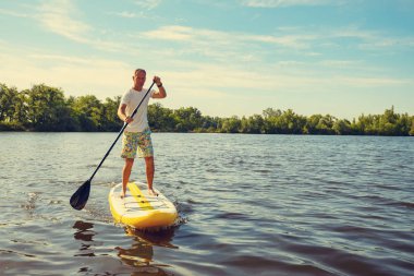 Neşeli adam Sup kurulu büyük nehir bir güneşli sabah bir turkuaz gökyüzü arka plan üzerinde eğitiyor. Yukarı raket yatılı - stand harika etkin açık rekreasyon. 