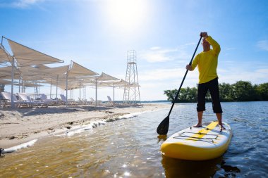 Neşeli adam bir Sup kurulu büyük nehir plaj ve zevk yaşamı boyunca içinde yola çıkacak. Yukarı raket yatılı - stand harika etkin açık rekreasyon. Geniş açı, arkadan.