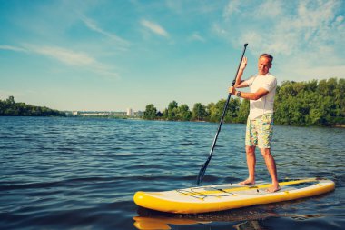 Neşeli adam Sup kurulu büyük nehir bir güneşli sabah bir turkuaz gökyüzü arka plan üzerinde eğitiyor. Yukarı raket yatılı - stand harika etkin açık rekreasyon.