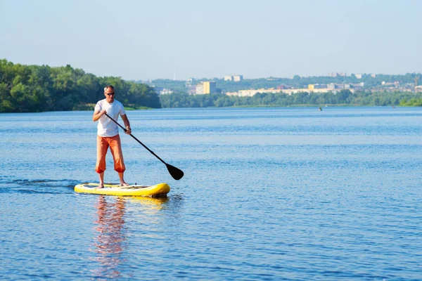 Man sails on a SUP board in large river on the cityscape background. Stand up paddle boarding - awesome active outdoor recreation.