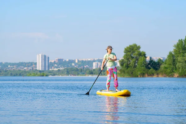 Woman sails on a SUP board in large river on the cityscape background. Stand up paddle boarding - awesome active outdoor recreation.