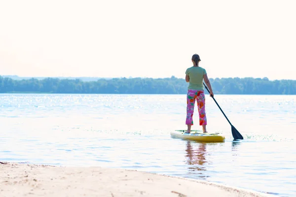 Woman sails on a SUP board in large river on a sunny morning. Stand up paddle boarding - awesome active outdoor recreation. Back view.