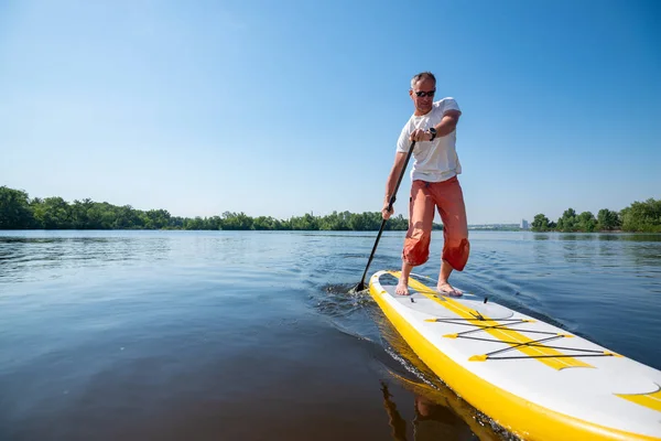 Joyful man in sunglasses sails on a SUP board in large river and enjoying life. Stand up paddle boarding - awesome active outdoor recreation. Wide angle.