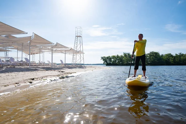 Joyful man sails on a SUP board in large river along the beach and enjoying life. Stand up paddle boarding - awesome active outdoor recreation. Wide angle, backlight.