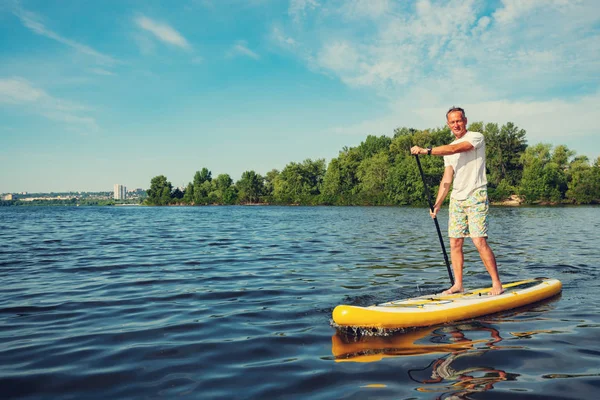 うれしそうな男は ターコイズ ブルーの空を背景に晴れた朝に大きな川で Sup ボードをトレーニングです スタンド アップ パドルボーディング 素晴らしいアクティブな野外レクリエーション — ストック写真