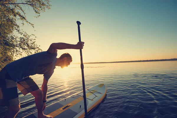 Man sails on a SUP board in a rays of rising sun. Stand up paddle boarding - awesome active outdoor recreation. Backlight, wide angle.