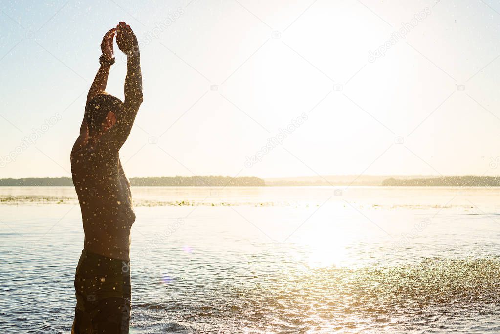 Joyful guy raises her hands with splashes of water in the rays of the rising sun - beach holidays. Back view, backlight.