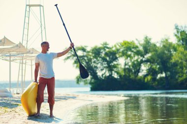 Neşeli adam standları Sup kurulu ile sahilde güneşli sabah ve zevk yaşam ile memnun. Yukarı raket yatılı - stand harika etkin açık rekreasyon.