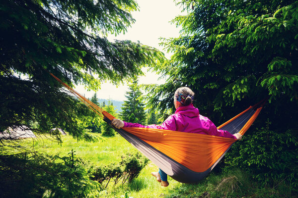 Adventurer female relaxes in hammock on the green mountain meadow among fir trees and admires view. Epic travel in the mountains. Back view.