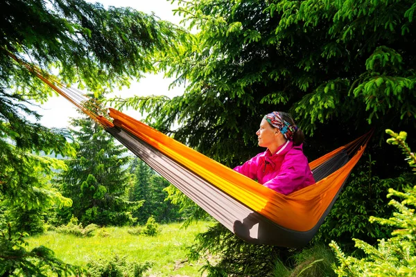 Adventurer female relaxes in hammock on the green mountain meadow among fir trees and admires view. Epic travel in the mountains.