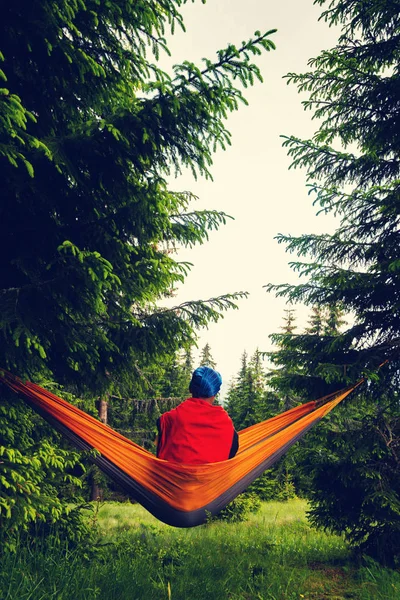 Adventurer relaxes in hammock on the green mountain meadow among fir trees and admires view. Epic travel in the mountains. Back view.