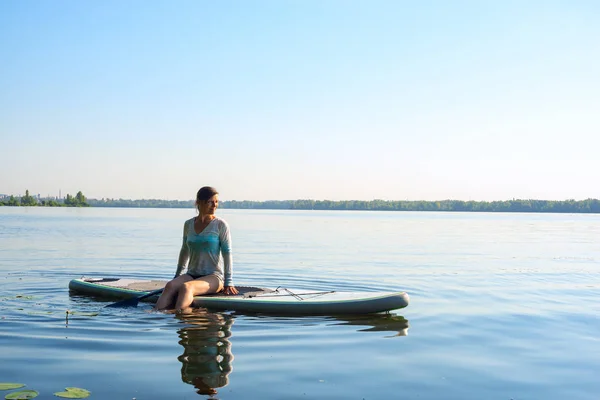 Happy female relaxing on a SUP board and enjoying life during sunny morning on a large river. Stand up paddle boarding - awesome active recreation in nature. Backlight.
