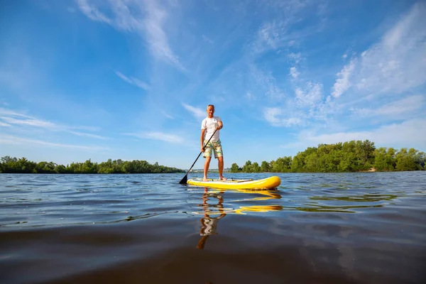 Joyful man paddling on a SUP board on large river and enjoying life, against the background of blue sky. Stand up paddle boarding - awesome active outdoor recreation. Wide angle.