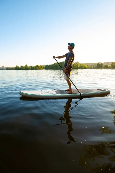 Guy paddling on a SUP board on large river during sunny evening and having fun. Stand up paddle boarding - awesome active outdoor recreation.