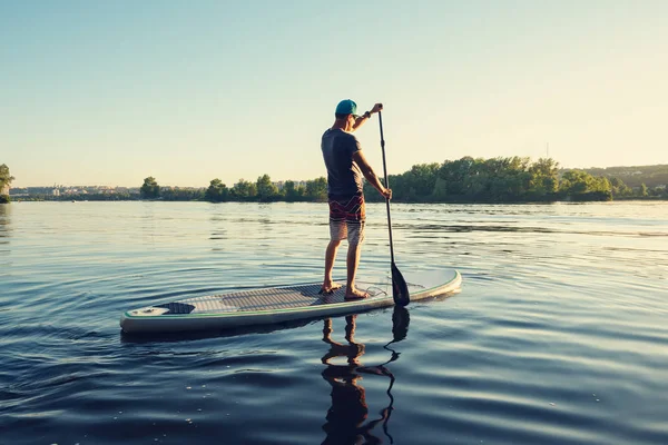 Guy paddling on a SUP board on large river during sunny evening. Stand up paddle boarding - awesome active outdoor recreation. Back view.