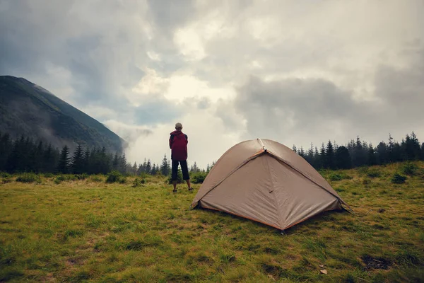 Adventurer relaxes in the camp on a green meadow and admires the mountain ranges in the fog after the storm. Epic travel in the mountains. Back view.