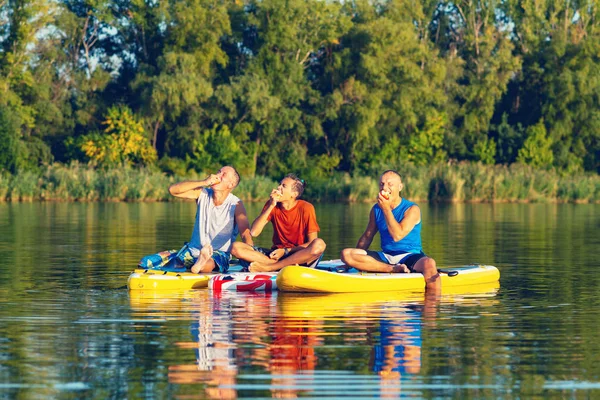 Joyful friends, a SUP surfers relax, eat apples and having fun during rest in nature. Stand up paddle boarding - awesome active outdoor recreation.