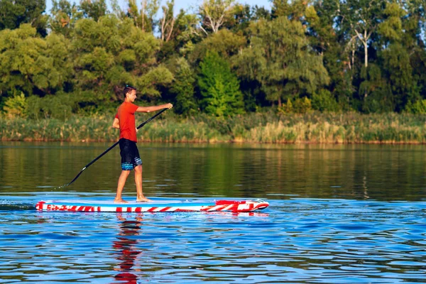 Happy guy, teenager paddling on a SUP board on large river and enjoying life. Stand up paddle boarding - awesome active outdoor recreation. Side view.