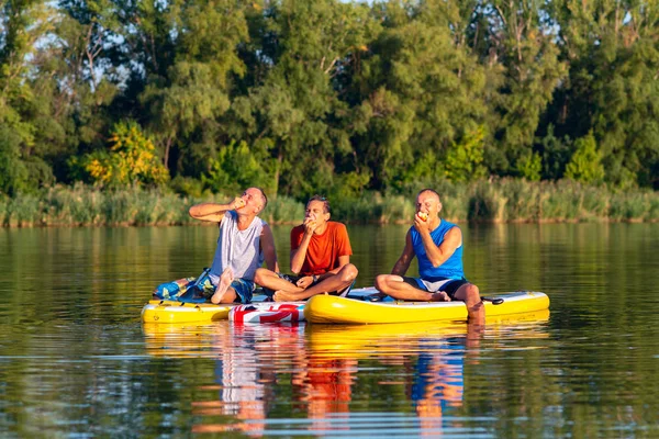 Joyful friends, a SUP surfers relax, eat apples and having fun during rest in nature. Stand up paddle boarding - awesome active outdoor recreation.