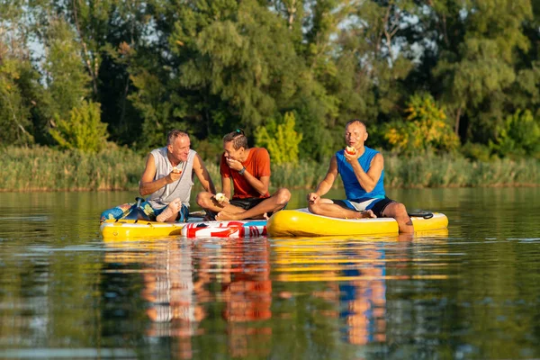 Happy friends, a SUP surfers relax, eat apples and having fun during rest in nature. Stand up paddle boarding - awesome active outdoor recreation.