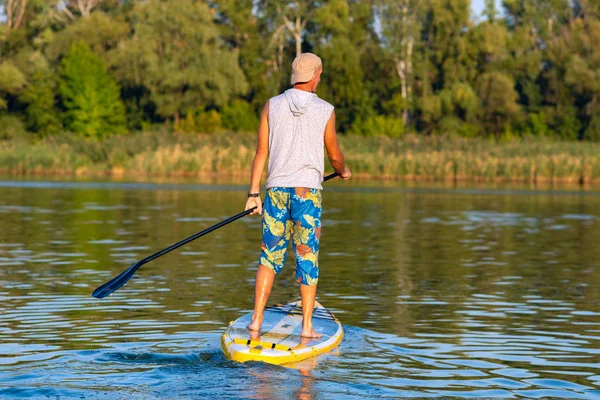 Happy man paddling on a SUP board on large river and enjoying life. Stand up paddle boarding - awesome active outdoor recreation. Back view.