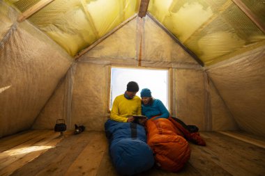 İki mutlu gezginler bir pencere yanında uyku tulumları içinde oturan ve bir tablet pc kullanarak eski dağ kulübede, dinlenme. Harika bir macera içinde kış ekilmemiş boş arazi.