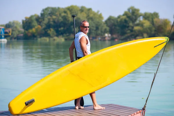 Sporty man stands on the beach with a SUP board is delighted with the sunny morning and smiling. Stand up paddle boarding - awesome active outdoor recreation.