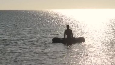 Adam bir şişme şişme deniz meditasyon, yoga sallanan dalgalar üzerinde güneşli bir akşamı uygulamak. Arkadan aydınlatmalı.