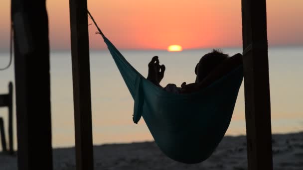 男は彼の前の海で夕日を眺めバンガロー ベランダのハンモックでリラックスします ビーチの素晴らしい夕日 背面図 — ストック動画