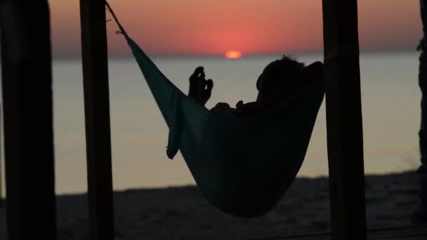 男人在平房凉廊的吊床上放松 在他面前欣赏大海的夕阳 海滩上令人惊叹的日落 后视图 — 图库视频影像
