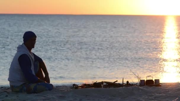海岸で焚き火の隣に旅行者の座っている瞑想し 海の向こうの夕日を賞賛します ビーチでの素晴らしいサンセット — ストック動画