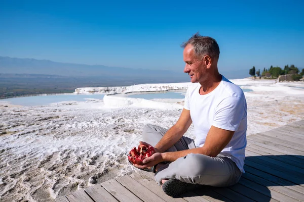 Happy traveler relaxes with a ripe pomegranate in his hands on the background of snow-white hills  - Pamukkale, Turkey tourist landmark.