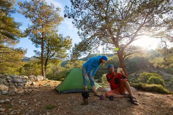 Couple of happy travelers is drinking coffee in the mountains among lush pines at sunny morning and using tablet pc - camp during adventure travel along Lycian way, Turkey. Backlight, wide angle.