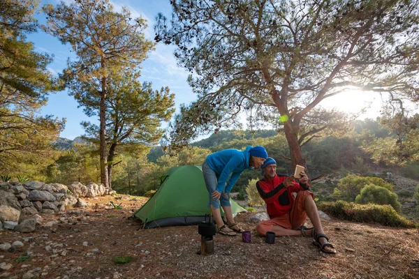 Couple of happy travelers is drinking coffee in the mountains among lush pines at sunny morning and using tablet pc - camp during adventure travel along Lycian way, Turkey. Backlight, wide angle.