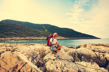 Seyahat çantasını için bağlı bir taşınabilir güneş pili ile kayalık sahil üzerinde oturan ve tablet pc - macera seyahat boyunca Likya yolu, Türkiye'de kullanıyor. Arka ışık.