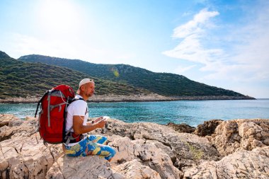 Mutlu traveler sırt için bağlı bir taşınabilir güneş pili ile kayalık sahil üzerinde oturuyor ve kullanarak tablet pc - macera seyahat boyunca Likya yolu, Türkiye'de. Arka ışık.